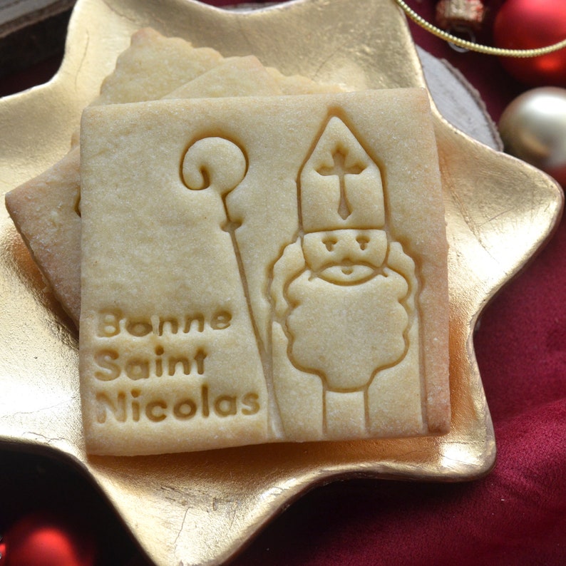 May include: Square shortbread biscuits with an embossed image of Saint Nicholas holding a staff, and the words "Bonne Saint Nicolas". The biscuits are light brown and rest on a gold star-shaped plate.