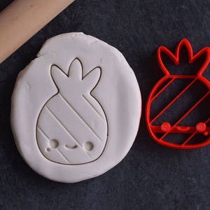 May include: A red pineapple-shaped cookie cutter next to a piece of dough with a pineapple imprint. A wooden rolling pin is in the background. The pineapple design has a smiling face and diagonal lines.