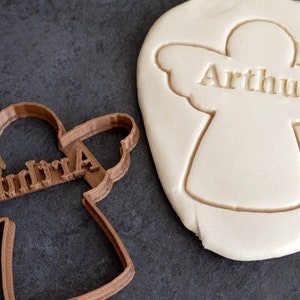 Könnte beinhalten: Ein brauner Plastik-Engel-Ausstecher mit dem Namen "Arthur" in Prägung. Im Hintergrund ist ein weißer Fondant-Engel-Keks mit dem Namen "Arthur" in Prägung zu sehen.