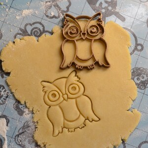 May include: A close-up of a brown cookie cutter in the shape of an owl, pressed into a round piece of cookie dough on a blue and white patterned surface.