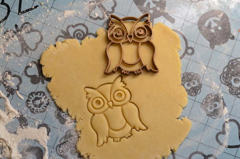 May include: A close-up of a brown cookie cutter in the shape of an owl, pressed into a round piece of cookie dough on a blue and white patterned surface.