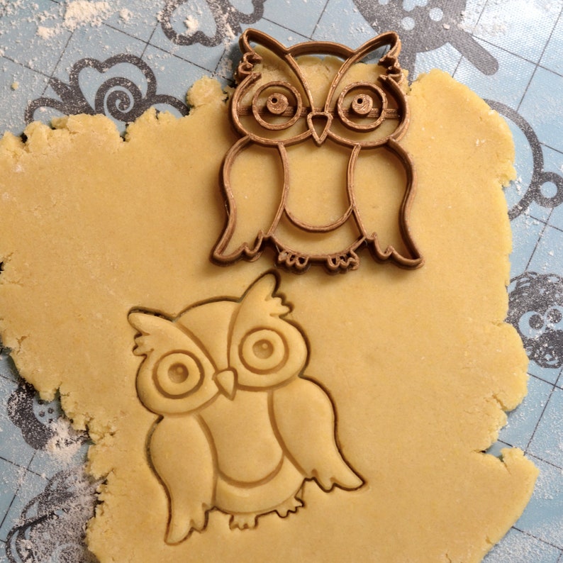 May include: Two brown metal owl cookie cutters on a light brown cookie dough background. The larger owl is on top of the smaller owl.