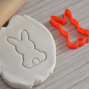 Puede incluir: Una masa de galletas blanca con una impresi&oacute;n de cortador de galletas en forma de conejo. El cortador es rojo y se muestra al lado de la masa.
