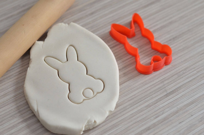 Puede incluir: Una masa de galletas blanca con una impresi&oacute;n de cortador de galletas en forma de conejo. El cortador es rojo y se muestra al lado de la masa.
