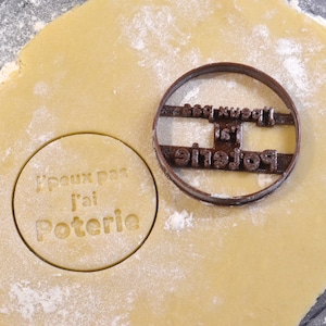 Puede incluir: Un cortador de galletas de color marrón con el texto "J'peux pas j'ai Poterie". El cortador de galletas está sobre una hoja de masa.