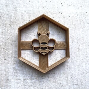 May include: Gold-coloured plastic cookie cutter shaped like a hexagon with a bee design in the centre.