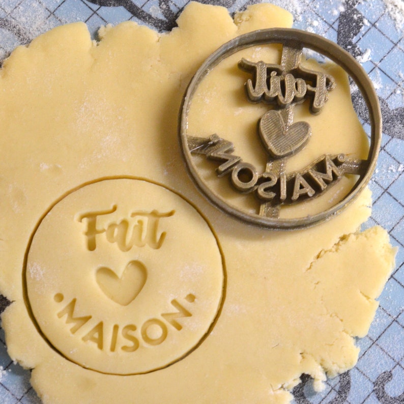 Puede incluir: Un primer plano de un cortador de galletas con las palabras "fait maison" y un dise&ntilde;o de coraz&oacute;n. El cortador est&aacute; encima de una masa de galletas.