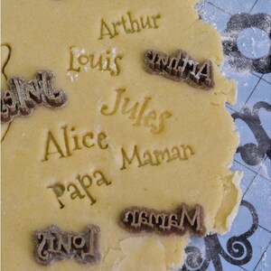 May include: A close-up of a sheet of cookie dough with various names pressed into it, including "Arthur", "Louis", "Alice", "Papa", "Jules", "Maman", and "Grandma".