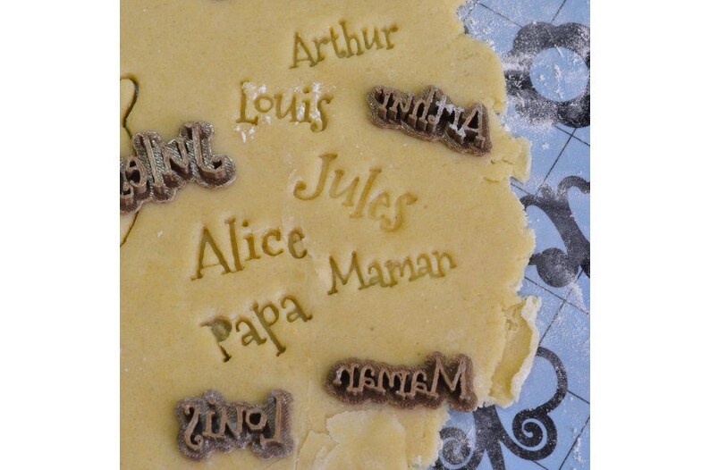 May include: A close-up of a sheet of cookie dough with various names pressed into it, including "Arthur", "Louis", "Alice", "Papa", "Jules", "Maman", and "Grandma".
