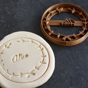 May include: A brown plastic cookie cutter in the shape of a wreath with the word "Alice" inside. The cutter is on a gray surface next to a white fondant cookie with the same design.