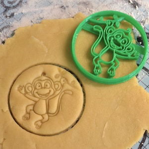 May include: A green plastic monkey-shaped cookie cutter next to a cut-out cookie. The cookie cutter is round and features a monkey hanging from a branch. The cut-out cookie is also round and shows the monkey design.