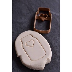 May include: A brown plastic cookie cutter in the shape of a mason jar with a heart cut out. The cookie cutter is on a gray surface with a white cookie dough cutout of the same shape.