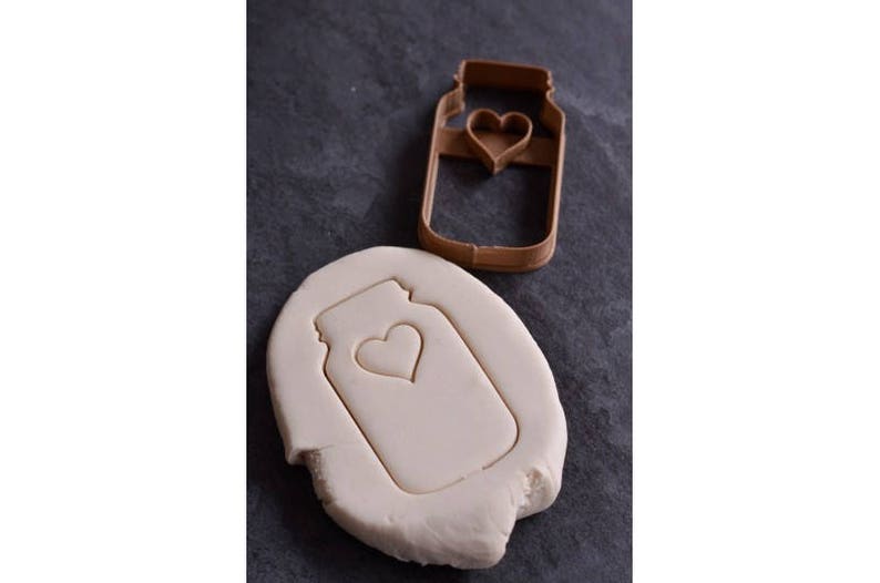 May include: A brown plastic cookie cutter in the shape of a mason jar with a heart cut out. The cookie cutter is on a gray surface with a white cookie dough cutout of the same shape.