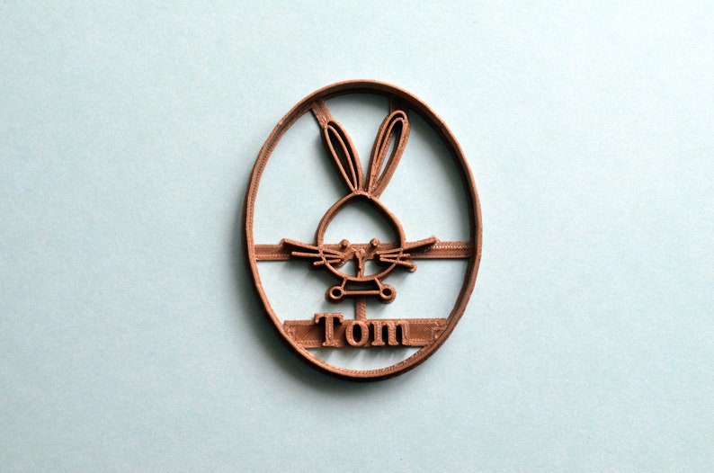 May include: Brown plastic cookie cutter shaped like an oval with a bunny rabbit inside and the name "Tom" underneath.