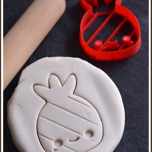 May include: A red cookie cutter in the shape of a cartoon fruit, next to a rolled-out dough with the same shape pressed into it. A wooden rolling pin is in the background. The website www.laboiteacookies.com is visible.
