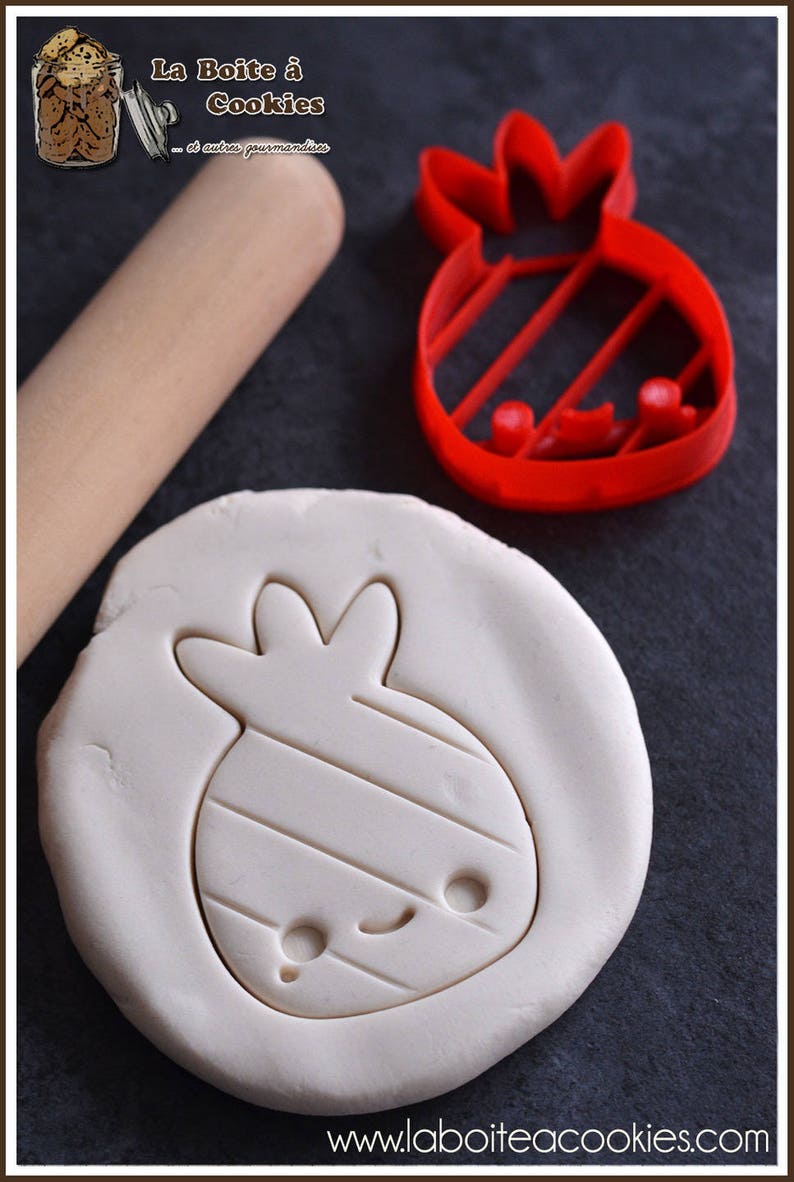 May include: A red cookie cutter in the shape of a cartoon fruit, next to a rolled-out dough with the same shape pressed into it. A wooden rolling pin is in the background. The website www.laboiteacookies.com is visible.
