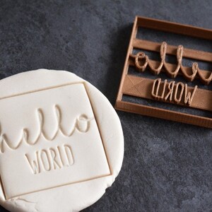 Op de afbeelding: Een bruine plastic koekjesvormer met de woorden "hello world" in een cursief schrift. De koekjesvormer ligt op een grijs oppervlak en is gebruikt om dezelfde woorden in een stuk wit deeg te stempelen.