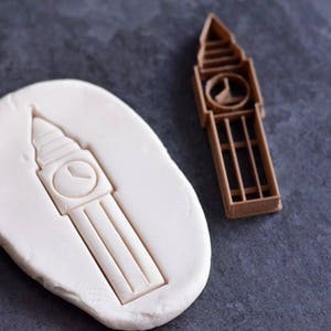 May include: A brown plastic cookie cutter shaped like the Big Ben clock tower, positioned next to a white, oval-shaped piece of dough that has been imprinted with the same clock tower design, showing the raised details of the tower structure.
