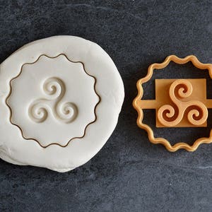 Op de afbeelding: Een witte koekdeeg met een bruine omtrek van een triskelion-symbool. Een bruine plastic koekjesvormer met een triskelion-symbool.