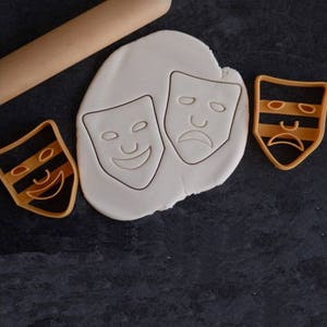 Peut inclure: Trois emportes-pièces en forme de masques théâtraux. L'un est un masque joyeux, l'autre un masque triste et le dernier un masque neutre. Les emportes-pièces sont sur une surface blanche avec un rouleau à pâtisserie en arrière-plan.