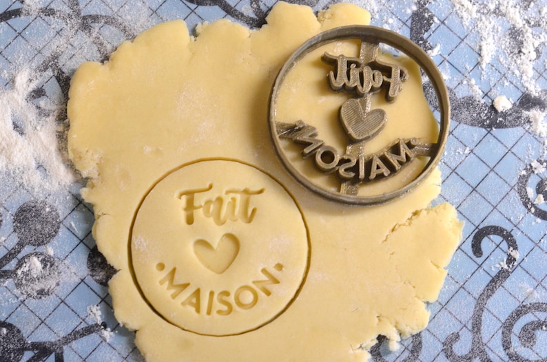 Puede incluir: Un primer plano de un cortador de galletas con el texto "Petit Maison" y un coraz&oacute;n, junto a una masa de galletas con el texto "Fait Maison" y un coraz&oacute;n.