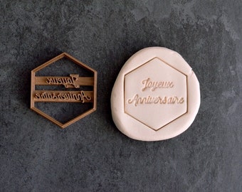 May include: A brown plastic cookie cutter in the shape of a hexagon with the words "Joyeux Anniversaire" embossed on it. The cutter is next to a white piece of fondant with the same words embossed on it.