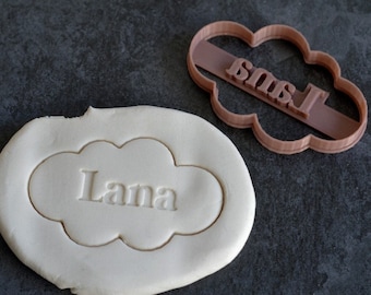 May include: A brown plastic cookie cutter in the shape of a cloud with the name "Lana" inside. The cutter is next to a piece of white dough with the name "Lana" imprinted in it.