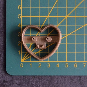 Pode incluir: Um cortador de biscoitos de pl&aacute;stico marrom em forma de cora&ccedil;&atilde;o com um rosto sorridente. O cortador tem dois pequenos olhos pretos e uma boca curva.