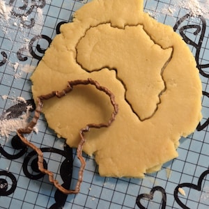 Op de afbeelding: Een koekjesvorm in de vorm van het Afrikaanse continent ligt naast een uitsparing in dezelfde vorm in een stuk deeg. Het deeg ligt op een blauw oppervlak met een decoratief patroon en bloem.