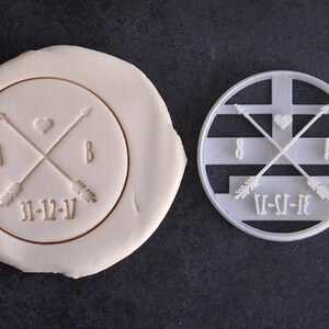 Puede incluir: Un cortador de galletas de plástico blanco con un diseño de dos flechas que se cruzan en el medio con un corazón en la intersección. Las flechas tienen plumas y el texto "A & B 31-12-V" está escrito debajo de las flechas.