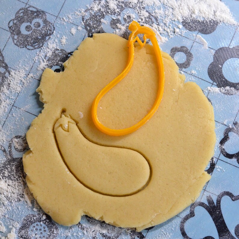 May include: A yellow plastic cookie cutter in the shape of an eggplant is on top of a round piece of cookie dough. The cookie dough is on a blue and white patterned surface.