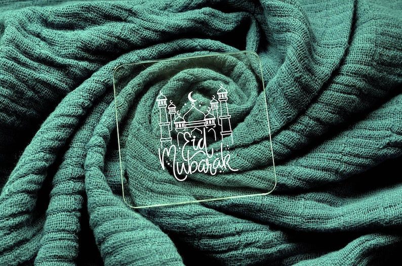 May include: A clear glass square with a white outline of a mosque and the text "Eid Mubarak" on a teal fabric background.