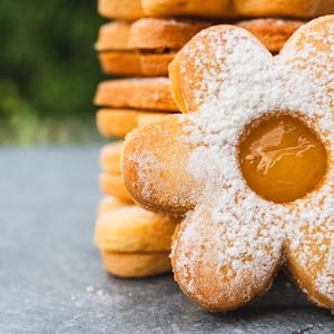 Può includere: Primo piano di biscotti a forma di fiore spolverati di zucchero a velo, con un ripieno giallo al centro. Una pila di biscotti simili è visibile sullo sfondo, suggerendo un tema di prodotti da forno.