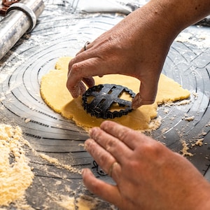 May include: A person using a black cookie cutter to cut out a shape from a sheet of yellow dough on a gray surface with white flour scattered around.