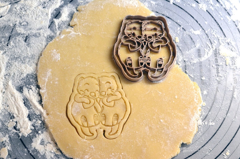 May include: A brown plastic cookie cutter in the shape of two hugging animals. The cutter is on top of a round piece of dough, ready to be used to make cookies.