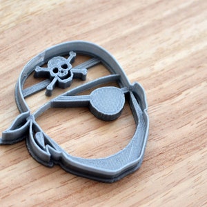 Könnte beinhalten: Ein grauer Plastik-Ausstecher in Form eines Piratenkopfes mit Augenklappe und Totenkopf mit Knochen auf einem Bandana.