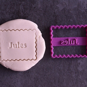 May include: A purple plastic cookie cutter in the shape of a rectangle with scalloped edges. The cutter has the word "Jules" embossed on it. A white fondant cookie is shown with the cutter pressed into it.