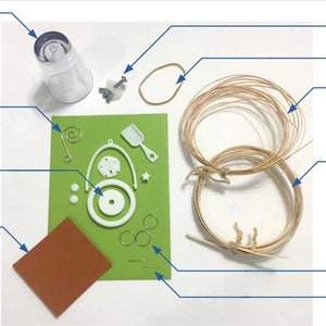 May include: A set of craft supplies for making a resin sand pail ornament. The set includes a clear plastic mold, a brown rubber band, a white resin sand pail set, a green sheet of paper with white resin pieces, a brown piece of sandpaper, two brown reeds, and three silver rings.
