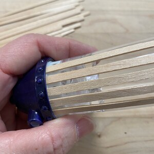 May include: A close-up of a blue and clear diffuser with wooden sticks inserted into the top. The diffuser is being held by a hand.