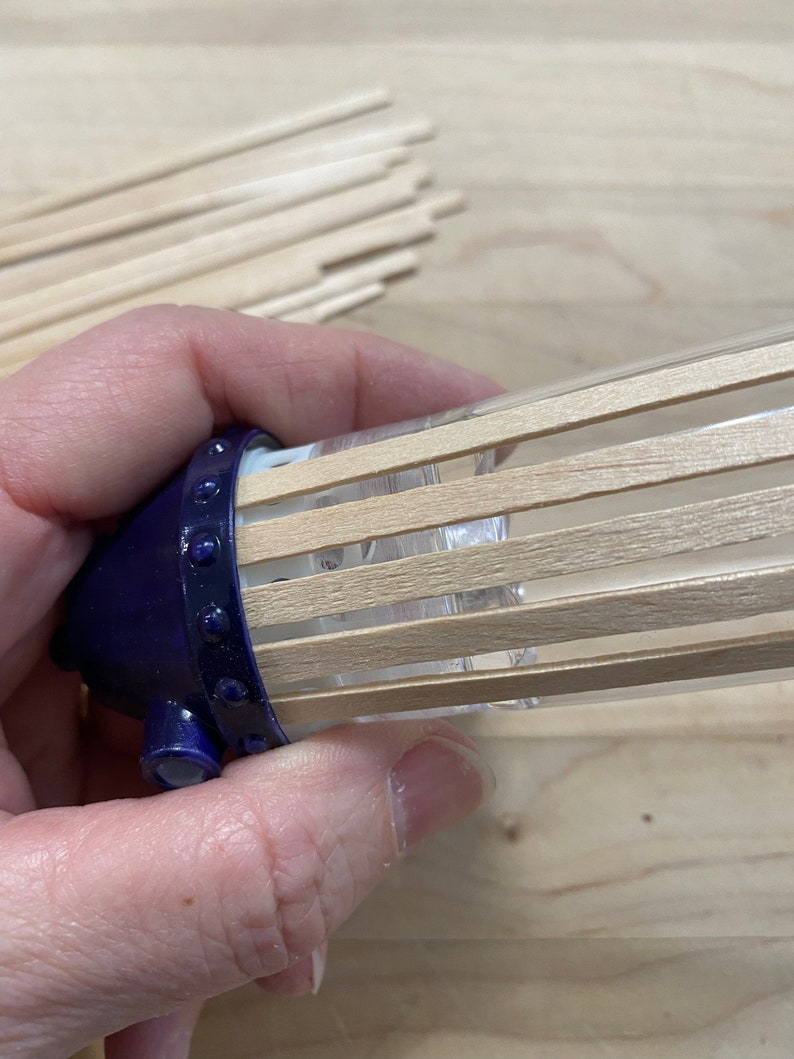 May include: A close-up of a blue and clear diffuser with wooden sticks inserted into the top. The diffuser is being held by a hand.