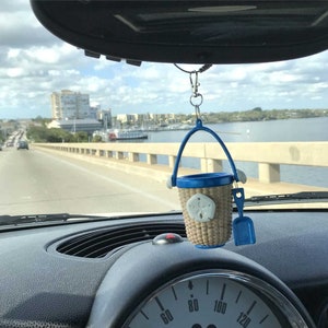 May include: A blue and tan miniature beach pail and shovel hanging from a rearview mirror in a car. The pail has a blue handle and a tan woven body with a white seashell design. The shovel is blue plastic.
