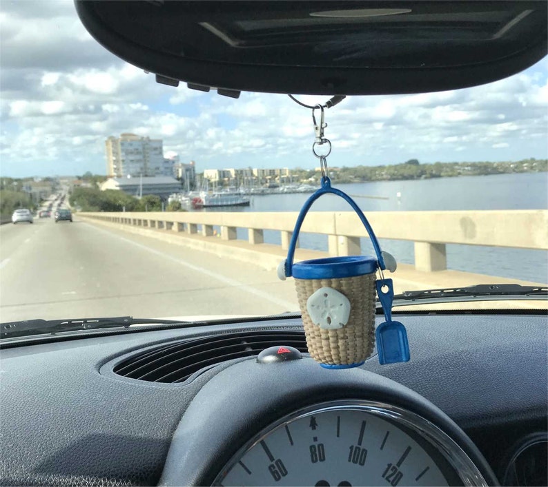 May include: A blue and tan miniature beach pail and shovel hanging from a rearview mirror in a car. The pail has a blue handle and a tan woven body with a white seashell design. The shovel is blue plastic.