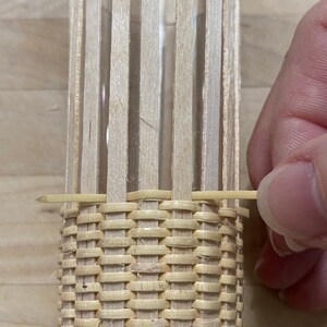 May include: A close-up of a woven basket being made around a glass cylinder. The basket is made of thin, light brown strips of wood that are woven together in a tight pattern. The cylinder is visible through the woven strips.
