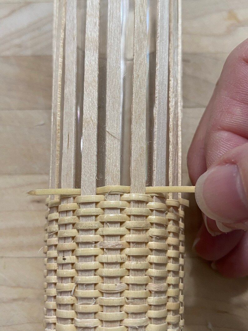 May include: A close-up of a woven basket being made around a glass cylinder. The basket is made of thin, light brown strips of wood that are woven together in a tight pattern. The cylinder is visible through the woven strips.
