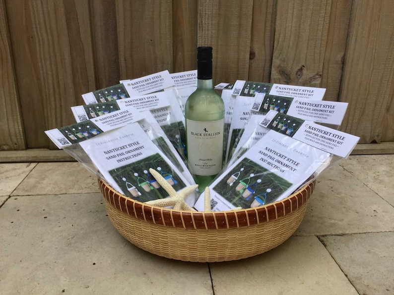 May include: A wicker basket filled with multiple packages of Nantucket Style Sand Pail Ornament Kits. The kits include instructions and all the materials needed to create a sand pail ornament. A bottle of white wine is also included in the basket.