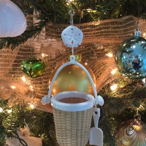 May include: A white wicker basket ornament with a white plastic shovel hanging from a gold and white ornament. The ornament is hanging on a Christmas tree decorated with burlap, lights, and other ornaments.