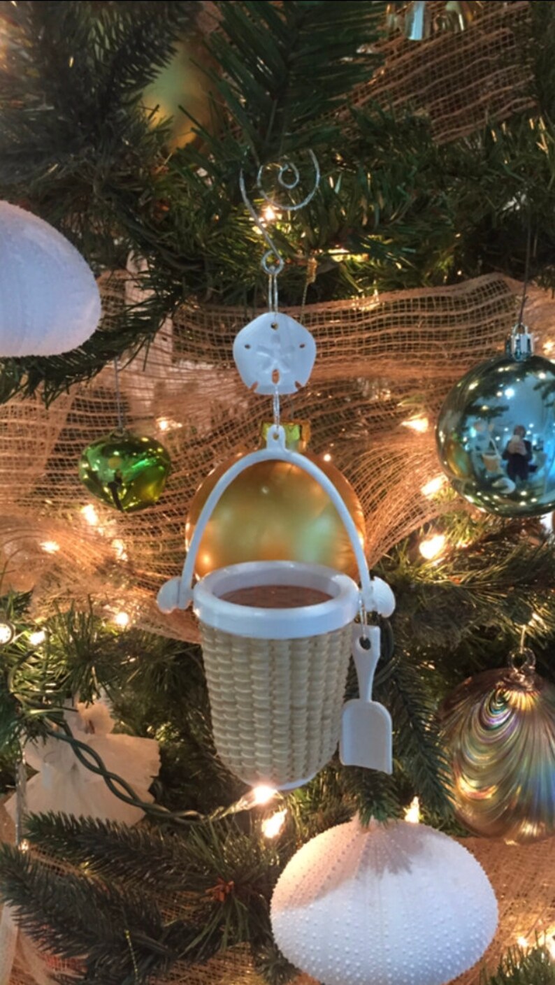 May include: A white wicker basket ornament with a white plastic shovel hanging from a gold and white ornament. The ornament is hanging on a Christmas tree decorated with burlap, lights, and other ornaments.