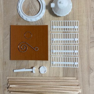 May include: A kit for building a model of a lighthouse. The kit includes white plastic pieces, wooden dowels, a brown felt sheet, and a metal wire.
