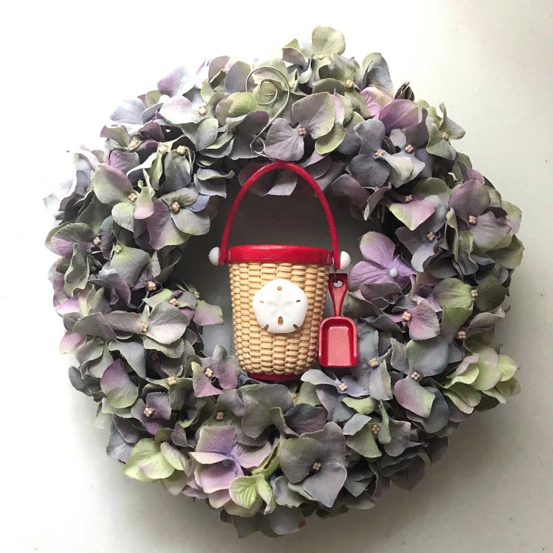 May include: A purple and green floral wreath with a red and white plastic bucket and shovel in the center. The bucket has a seashell design.