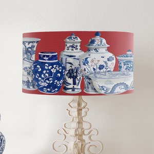 May include: A white table lamp with a red lampshade featuring a pattern of blue and white porcelain vases. The lampshade has a white trim around the top and bottom. The lamp base is white and has a decorative design.